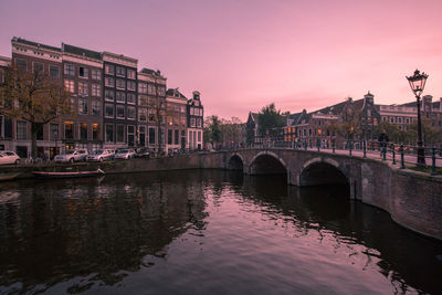 Bridge over river by buildings against sky at sunset