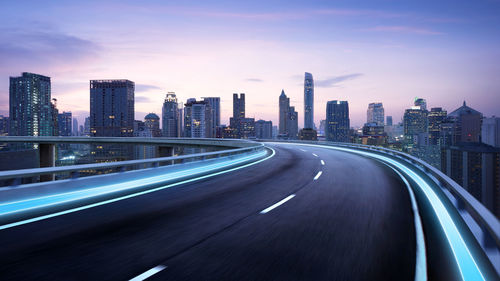 Light trails on elevated road by illuminated buildings against sky in city