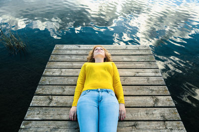 High angle of tranquil female in casual wear lying on wooden pier near lake and enjoying silence on summer day