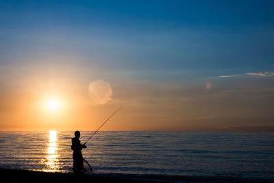 Silhouette man fishing at beach against sky during sunset