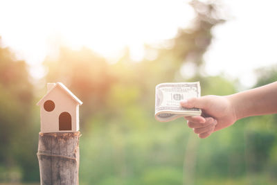 Close-up of hand holding paper currency with model house on wooden post against sky