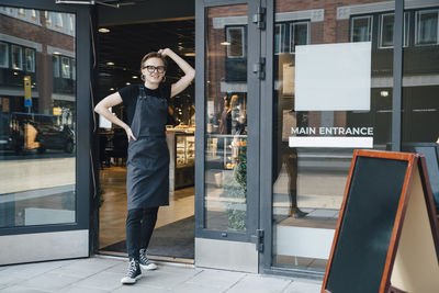 Full length portrait of smiling young owner standing at cafe entrance