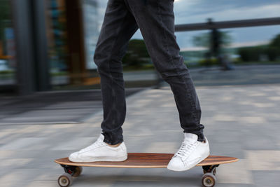 Cropped shot of young man in black jeans and white sneakers riding on longboard