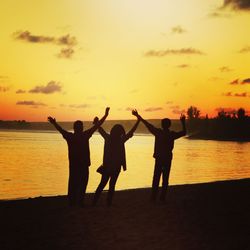 Silhouette friends standing with raised hands at beach against orange sky during sunset