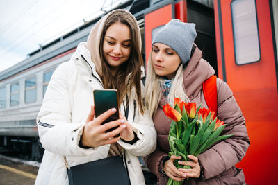 Two girls on the station platform with flowers happily looking into a smartphone