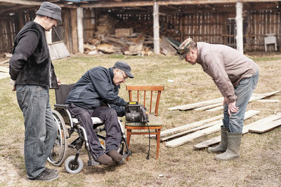 Elderly man  in wheelchair is repairing  old electric saw in his yard. his two friends watching 