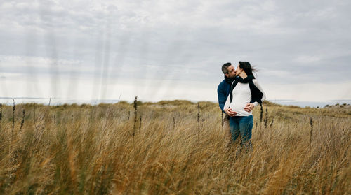 Couple kissing while standing on grassy field against cloudy sky