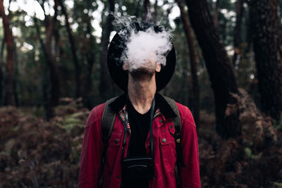 Hipster traveler in hat exhaling cloud of smoke standing along against dark trees of woods