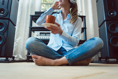 Low section of woman listening music while holding coffee cup on floor