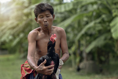 Shirtless man holding rooster while standing against trees