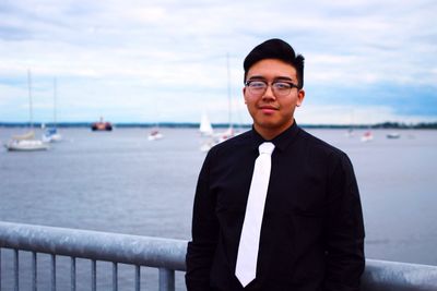 Portrait of smiling businessman standing by railing at harbor against cloudy sky