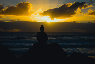 Rear view of silhouette man sitting at beach against cloudy sky during sunset