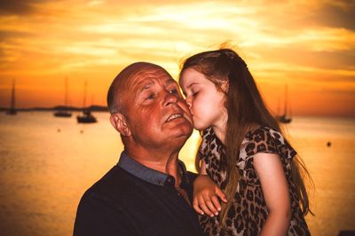 Girl kissing grandfather while standing at beach during sunset