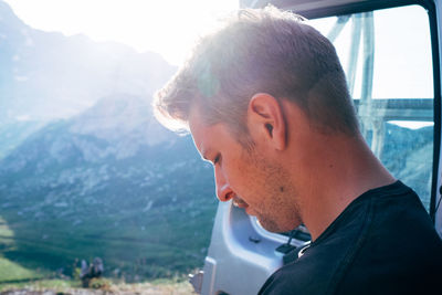 Close-up of young man looking down while standing by car door on mountain