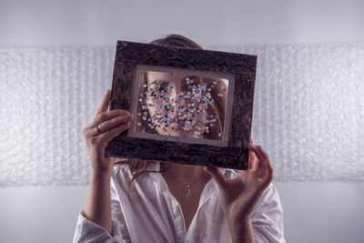 Woman covering face with picture frame at home