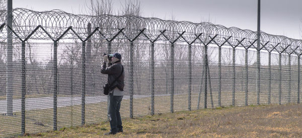 Side view of man photographing by chainlink fence on field