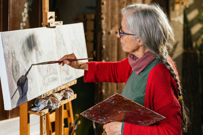 Side view of aged female artists in protective mask painting on canvas in backyard on sunny day in summer