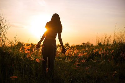 Young woman walking in spring field at sunset among fresh grass and touching yellow flowers.