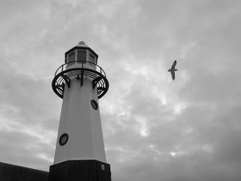 Low angle view of seagull flying by lighthouse against cloudy sky