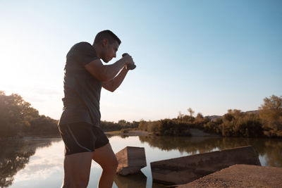 Man exercising while standing by lake against sky during sunset