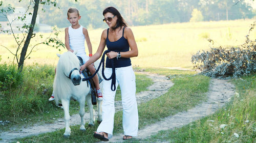 Mom and daughter are walking around the field, daughter is riding a pony, mother is holding a pony
