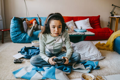 Woman playing video game while female friend using smart phone on sofa at home