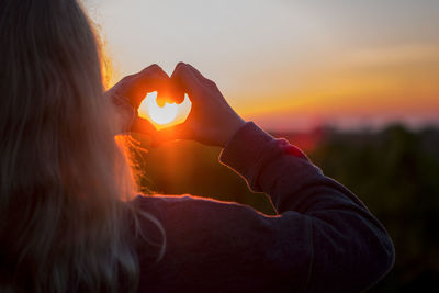 Rear view of woman making heart shape against sunset sky