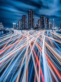 Light trails on city street by buildings against sky at night