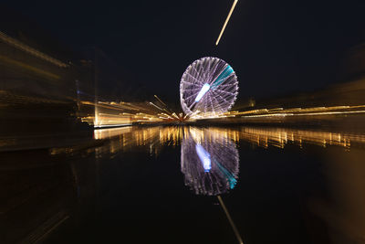 Illuminated ferris wheel by lake against sky at night