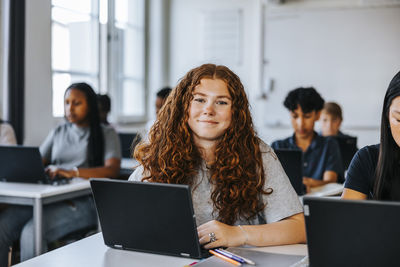 Portrait of smiling curly hair teenage student sitting with laptop near desk in classroom at school