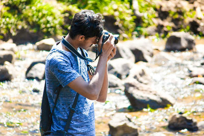 Young man photographing stream through camera at forest