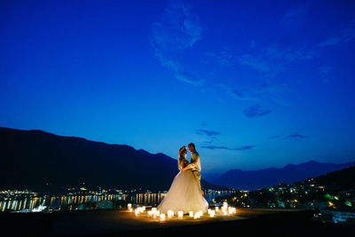 Woman standing by illuminated mountain against blue sky at dusk