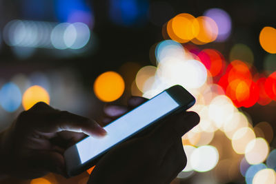 Cropped hands of person using phone against defocused lights at night