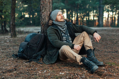 Young man looking away while sitting by tree trunk in forest