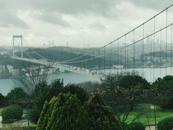 High angle view of suspension bridge in city against cloudy sky
