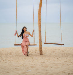 Full length of woman sitting on rope swing at beach against sky