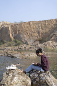 Man sitting on rock by mountain against sky