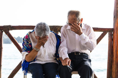 Senior couple covering face while sitting on bench with sea in background