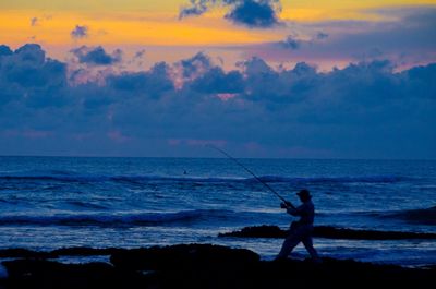 Silhouette man fishing at beach against sky during sunset