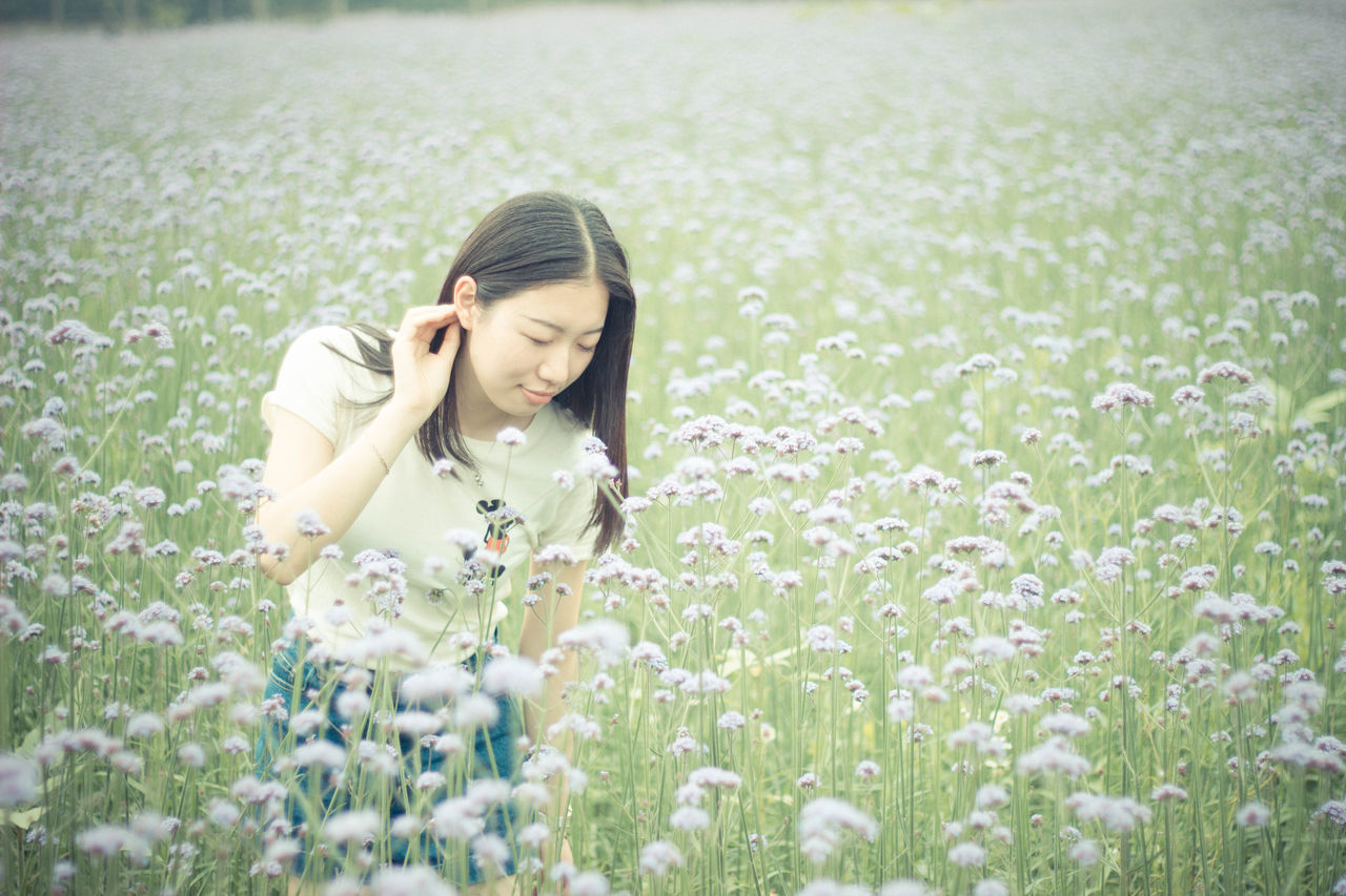 person, lifestyles, young adult, leisure activity, young women, casual clothing, smiling, grass, field, flower, looking at camera, portrait, standing, focus on foreground, plant, happiness, three quarter length, nature