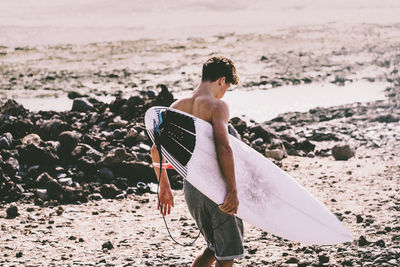 Shirtless boy holding surfboard while standing on beach