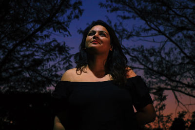 Portrait of young woman standing against trees at dusk