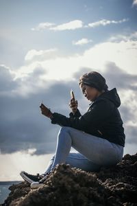 Side view of woman sitting on rock against sky