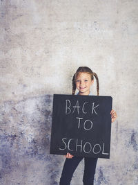 Portrait of smiling girl holding writing slate with text against wall