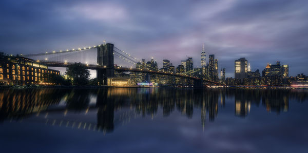 Illuminated bridge over river by buildings against sky at dusk