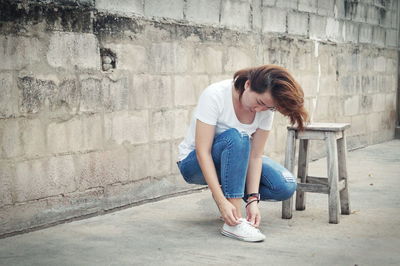Young woman tying shoes while crouching against wall