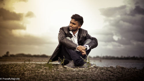 Young man looking away while sitting on land against sky