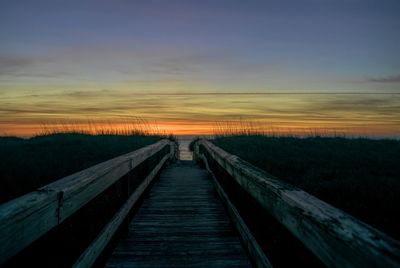 Boardwalk leading towards bridge against sky during sunset