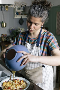 Woman pouring batter from bowl on chopped fruits in kitchen