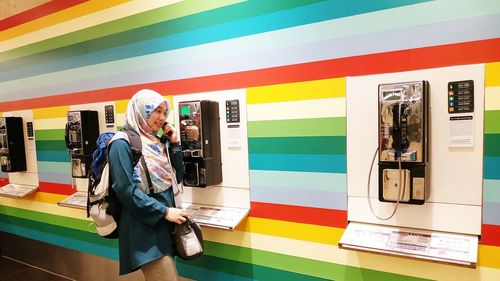 Smiling woman talking through pay phone mounted on multi colored wall